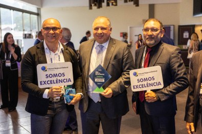 A imagem mostra um homem de terno cinza claro sorrindo e segurando um troféu azul do Prêmio CNJ ...