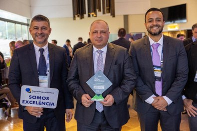 A imagem mostra um homem de terno cinza claro sorrindo e segurando um troféu azul do Prêmio CNJ ...