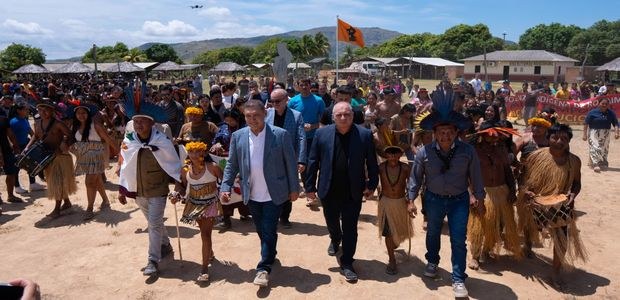 Em uma área aberta de terra batida, um grande grupo de pessoas participa de uma caminhada coleti...