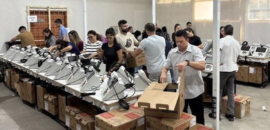 Ambiente interno com várias urnas eletrônicas dispostas sobre mesas compridas. Diversas pessoas ...