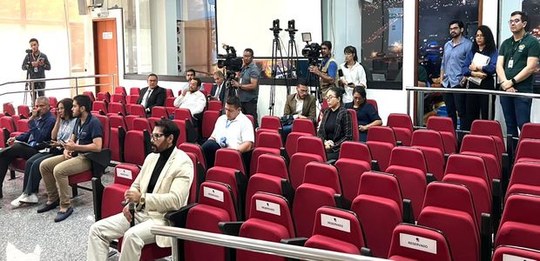 Auditório com várias fileiras de cadeiras vermelhas, algumas ocupadas por pessoas sentadas. Ao f...
