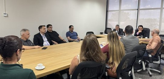 Sala de reuniões com uma mesa retangular de madeira cercada por várias pessoas sentadas. Os part...