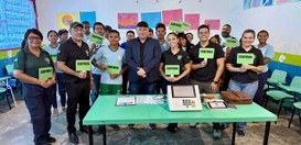 Foto em ambiente escolar mostrando um grupo de cerca de vinte pessoas reunidas atrás de uma mesa...