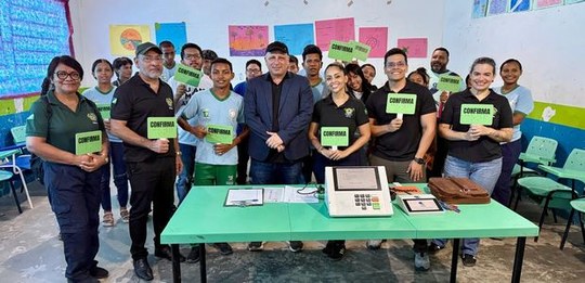 Foto em ambiente escolar mostrando um grupo de cerca de vinte pessoas reunidas atrás de uma mesa...