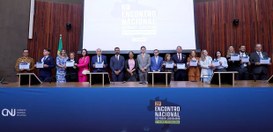 Foto de cerimônia institucional realizada em um auditório. Em um palco, várias pessoas estão ali...