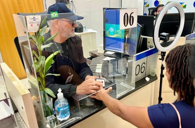 Atendimento em balcão de órgão público: duas pessoas interagem através de uma divisória de vidro...