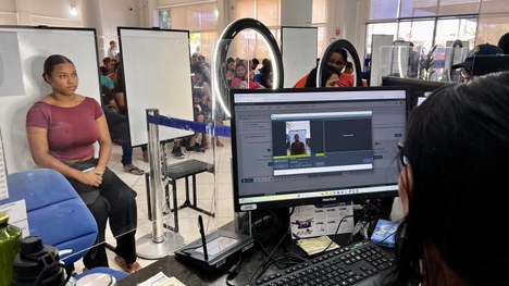 Atendimento em balcão de órgão público: duas pessoas interagem através de uma divisória de vidro...