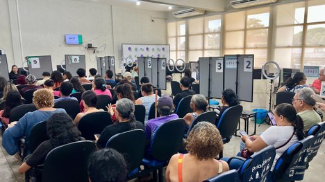 Atendimento em balcão de órgão público: duas pessoas interagem através de uma divisória de vidro...