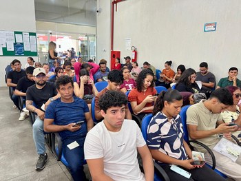 Atendimento em balcão de órgão público: duas pessoas interagem através de uma divisória de vidro...