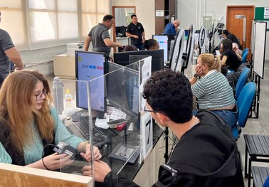 Atendimento em balcão de órgão público: duas pessoas interagem através de uma divisória de vidro...