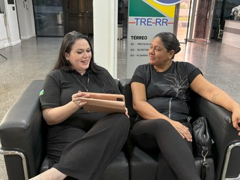 Duas mulheres sentadas em um sofá, vestindo camisa preta; uma está sorrindo e mostrando um table...