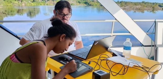 A imagem mostra duas pessoas sentadas em uma mesa amarela, que está posicionada em uma área exte...