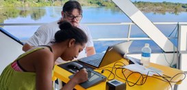 A imagem mostra duas pessoas sentadas em uma mesa amarela, que está posicionada em uma área exte...