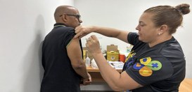 Em uma sala interna, uma pessoa aplica uma vacina no braço de outra pessoa. A pessoa vacinada es...
