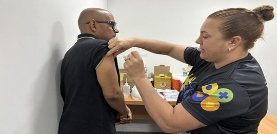 Em uma sala interna, uma pessoa aplica uma vacina no braço de outra pessoa. A pessoa vacinada es...