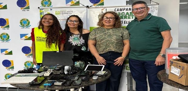 Quatro pessoas sorridentes posam atrás de uma mesa com vários eletrônicos usados, como celulares...