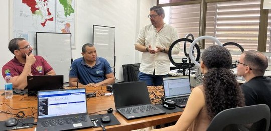 Em uma sala de reunião, várias pessoas estão sentadas ao redor de uma mesa com notebooks abertos...