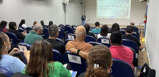 Público sentado em auditório acompanha apresentação. À frente, uma pessoa realiza palestra, com ...