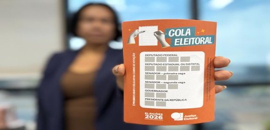 Pessoa segura, em primeiro plano, uma cola eleitoral nas cores laranja e branca, com espaços par...