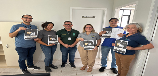 Foto em ambiente interno, com sete pessoas em pé, lado a lado, em um corredor de escritório. Tod...