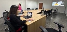 A cena mostra uma sala de reuniões moderna, com uma longa mesa de madeira clara em formato de “U...