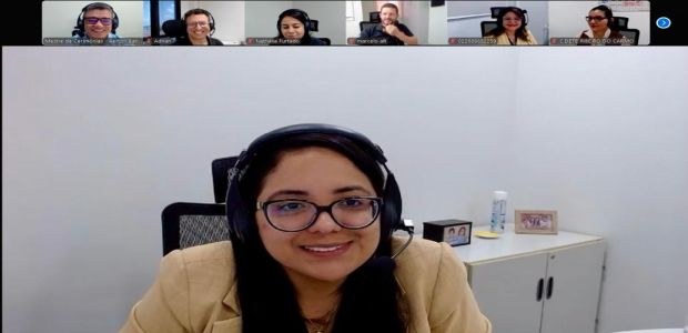 A imagem mostra uma mulher de óculos e fone de ouvido com microfone, sorrindo em frente à câmera...