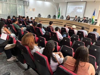 Auditório com várias pessoas sentadas em cadeiras vermelhas, voltadas para a frente, acompanhand...