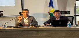 Mesa de reunião em ambiente institucional. Duas pessoas estão sentadas atrás da mesa, utilizando...