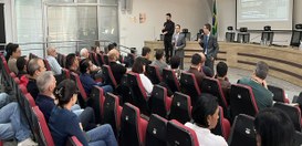 Fotografia de um auditório durante uma apresentação institucional. Pessoas estão sentadas em fil...