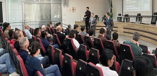 Fotografia de um auditório durante uma apresentação institucional. Pessoas estão sentadas em fil...