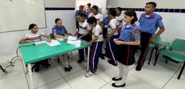 A foto mostra um grupo de estudantes uniformizados reunidos em uma sala de aula. Alguns estão se...