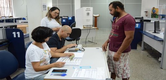 Ambiente interno de atendimento com mesas e divisórias, semelhante a uma seção eleitoral. Três a...