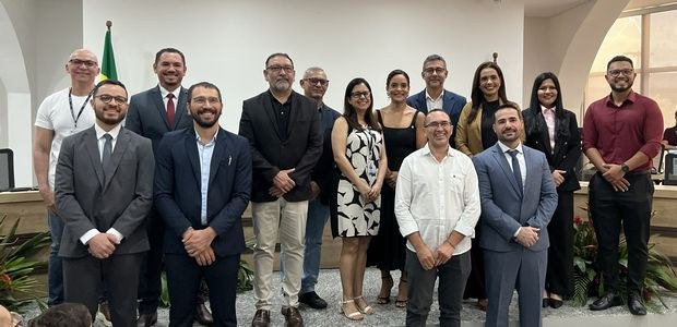 Foto de grupo em ambiente interno institucional. Treze pessoas estão posicionadas em duas fileir...
