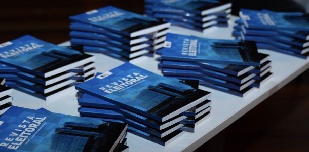 TRE-RR está recebendo artigos para segunda edição da Revista Eleitoral Uma mesa branca está cheia de pilhas de livros de capa azul. Na capa, lê-se “Revista Eleitoral” ...