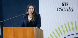 A imagem mostra uma mulher em pé atrás de um púlpito de madeira, falando ao microfone. Ao fundo,...