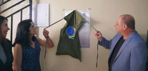 Em um ambiente interno, três pessoas estão diante de uma parede clara. Duas delas puxam cordões ...