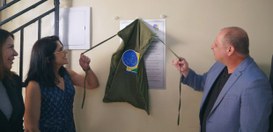 Em um ambiente interno, três pessoas estão diante de uma parede clara. Duas delas puxam cordões ...