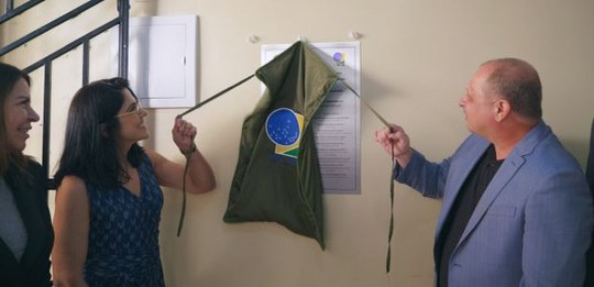 Em um ambiente interno, três pessoas estão diante de uma parede clara. Duas delas puxam cordões ...