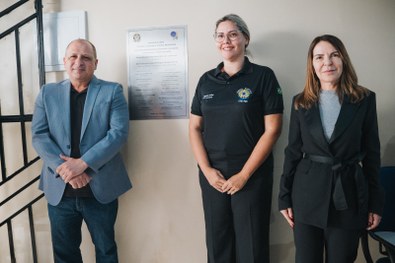 Em um ambiente interno, três pessoas estão posicionadas lado a lado em frente a uma parede clara...