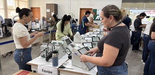 Ambiente de trabalho com várias pessoas organizando e manuseando urnas eletrônicas sobre mesas. ...