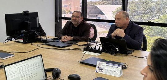 Foto de uma sala de reunião com uma mesa de madeira. Duas pessoas adultas estão sentadas lado a ...