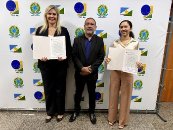 Foto em ambiente interno. Quatro pessoas estão em pé, alinhadas lado a lado, voltadas para a câm...