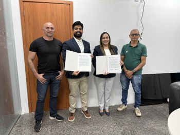 Foto em ambiente interno. Quatro pessoas estão em pé, alinhadas lado a lado, voltadas para a câm...