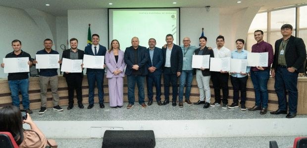 A imagem mostra um grupo de cerca de quinze pessoas em pé, alinhadas em um palco dentro de um au...