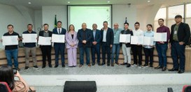 A imagem mostra um grupo de cerca de quinze pessoas em pé, alinhadas em um palco dentro de um au...