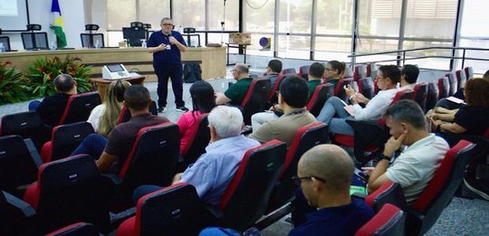 Foto de um auditório com várias pessoas sentadas em poltronas voltadas para a frente. Uma pessoa...