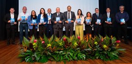 A imagem mostra um grupo de dez pessoas em pé sobre um palco, alinhadas lado a lado. Todas estão...