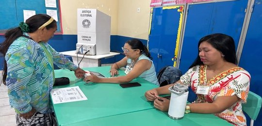 Em uma sala de votação, uma pessoa é atendida em uma mesa por duas servidoras da Justiça Eleitor...