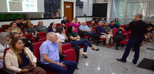 Sala de auditório com várias pessoas sentadas em cadeiras vermelhas, acompanhando uma palestra. ...