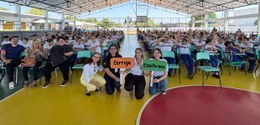 Fotografia de uma quadra poliesportiva coberta, repleta de estudantes uniformizados sentados em ...